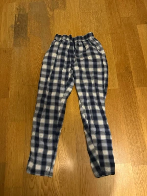 Rutiga mjukisbyxor med snörning - Säljer ett par rutiga mjukisbyxor i blått och vitt med elastisk midja och snörning. Byxorna är i mjukt bomullstyg och har en avslappnad passform, perfekta för chill dagar hemma.