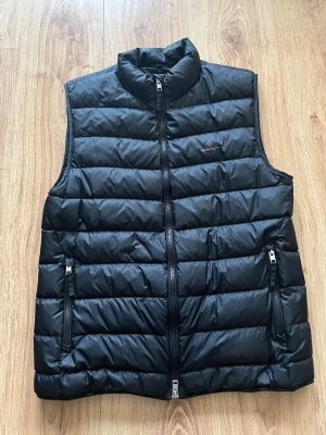 Svart dunväst från Gant - Snygg svart dunväst från Gant med quiltad design och hög krage. Västen har två dragkedjeförsedda fickor framtill och stängs med dragkedja. Perfekt att slänga över en hoodie eller tröja för extra värme.