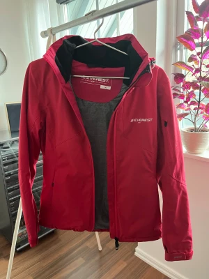 Röd/rosa Everest skidjacka med huva - Skidjacka från everest, storlek 36. Jackan är avsedd för vinterbruk och är tillverkad med material som ger skydd mot kyla. Den har huva som går att ta av. Dragkedja framtill samt fickor med dragkedja för förvaring.  Dragsko i midjan för enkel justering. Mycket bra skick och perfekt jacka att ha i skidbacken.
