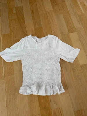 Vit blus med volangkant - Söt vit blus i bomull med korta ärmar och smockad framsida. Toppen har en volangkant nedtill som ger en extra detalj. Perfekt till jeans eller kjol för en fräsch look. Köpt på vinted
