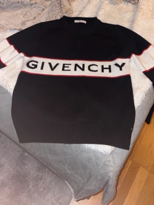Svart stickad tröja från Givenchy - Säljer en svart stickad tröja från Givenchy med vit och röd rand över bröstet och armarna. Stor Givenchy-logga framtill. Tröjan har rund hals och långa ärmar, samt ribbade muddar vid ärmslut och nederkant. Perfekt för dig som gillar exklusiva streetwear-plagg.