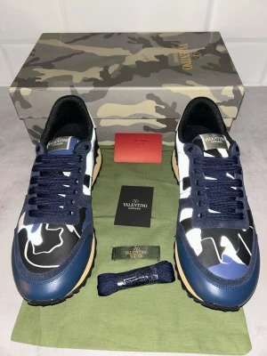Valentino Garavani Rockrunner sneakers - Säljer ett par Valentino Garavani Rockrunner sneakers med blå, svart och vit camouflage-mönster. Skorna har snörning, rund tå och platt sula med gummidetaljer. Materialmix av skinn och syntet. Kommer med originalkartong, extra skosnören och dustbag.