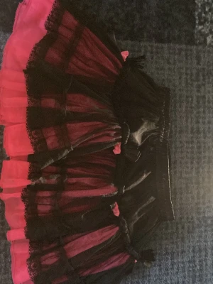 Svart och rosa volangkjol Poizen Industries - Kort kjol från Poizen Industries i svart och rosa med flera lager volanger och spetsdetaljer. Kjolen har elastisk midja och små rosetter som extra detalj. Perfekt för dig som gillar alternativ stil och vill sticka ut. Inget är fel med klädesplagget, bara passar inte min stil länge guys 🫰😴 storleken är xs fast har elastisk midja !! Så passar också som M (vilket jag är) 