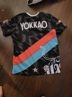 Yokkao t-shirt med stjärnor och tiger - Cool t-shirt från Yokkao med svart bas och stora stjärnor i bakgrunden. Framsidan har en vit tiger, röda och turkosa diagonala ränder samt tryckta stjärnor på ärmarna. Rund vit krage och tryckt logga på ryggen. Perfekt för dig som gillar sportig och unik stil.