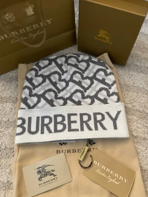 Vit och grå Burberry mössa - Säljer en snygg vit och grå mössa från Burberry med stort logotyp-tryck framtill och ett mönster av sammankopplade bokstäver. Mössan är stickad och har en bred uppvikt kant. Perfekt accessoar för att lyfta din vinterstil. Tillkommer bara med dustbag 