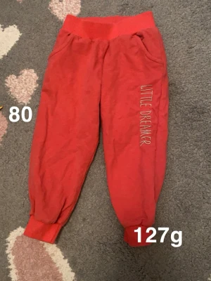 Rosa mjukisbyxor med text Little Dreamer - Säljer ett par rosa mjukisbyxor med bred resår i midjan och muddar vid bensluten. Byxorna har fickor och texten 'LITTLE DREAMER' broderad längs ena benet. Perfekta för chill dagar hemma eller till förskolan.🌸 Fint använt skick. Storlek 80