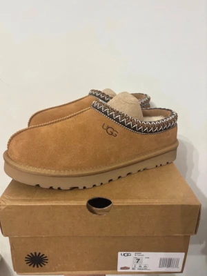 UGG  - Säljer ett par bruna slip-on boots från UGG med mjuk insida i päls och snyggt broderat mönster runt öppningen. Skorna har en rund tå och platt sula med grovt mönster undertill. Perfekta för kalla dagar och har en klassisk UGG-look.