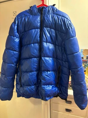 Blå pufferjacka med huva - Säljer en blå pufferjacka med huva och dragkedja framtill. Jackan har två sidofickor med dragkedja och quiltad design. Perfekt för kalla dagar och riktigt skön att bära. Passar dig som gillar en sportig och avslappnad stil.