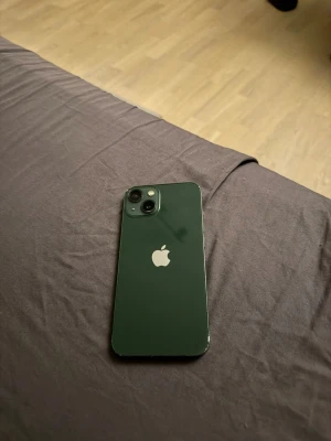 iPhone 13 Grön - Snygg grön iPhone 13 med dubbla kameror. Telefonen har några mindre repor och slitage på kanterna, men är i gott skick. Perfekt för dig som vill ha en kraftfull och pålitlig smartphone med modern design. Batterihälsa = 80-84%, 64 GB