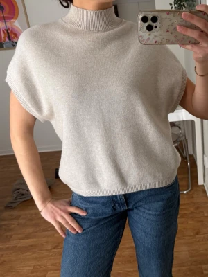 Beige ärmlös polotröja från Kappahl - Supermjuk beige polotröja från Kappahl i ärmlös modell. Tröjan har ribbad hög krage och är gjord i en blandning av viskos, polyester och polyamid. Perfekt att styla med jeans eller kjol för en clean och chill look. Strl S