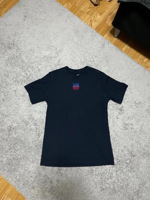 Svart Nike t-shirt med Barca-logga - Säljer en svart t-shirt från Nike med FC Barcelonas klubbmärke broderat på bröstet. Klassisk rund hals och korta ärmar, perfekt för dig som älskar fotboll och vill ha en clean look. Materialet känns mjukt och skönt mot huden.