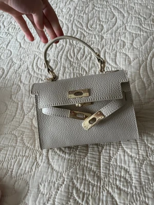 Vit handväska med gulddetaljer - Säljer en stilren vit cream handväska i strukturerat skinn med guldfärgade metalldetaljer och klassiskt vridlås. Väskan har ett topphandtag och medföljande axelrem för olika bärstilar. Perfekt storlek för det viktigaste och ger en clean look till din outfit. Genuin leather