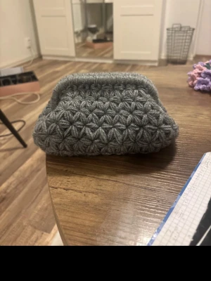 Grå virkad clutchväska  - Säljer en grå virkad kuvertväska med ett unikt blommigt mönster. Väskan har en mjuk och fluffig känsla, fyrkantig form och stängs med ett metallspänne. Perfekt för dig som gillar handgjorda accessoarer med personlig touch.