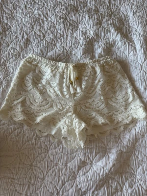 Vita spets-shorts från Hunkemöller - Säljer ett par vita spets-shorts från Hunkemöller i storlek S. De har elastisk midja med snörning och tofsar framtill. Materialet är en blandning av polyamid och polyester, vilket ger en mjuk och luftig känsla. Perfekta till sommaren för en romantisk och chill look.