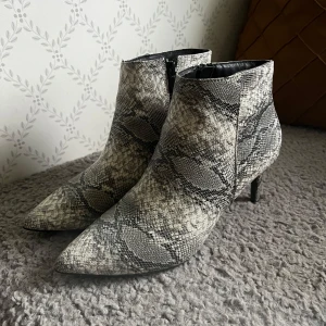 Klackar i ormskinn - Säljer ett par snygga stövletter med ormskinnsmönster i grått och svart. Skorna har spetsig tå, smal klack och dragkedja på sidan. Rätt så smala på bredden. Använda fåtal gånger så mycket fint skick! Ordpris. 499:-