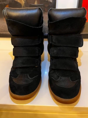 Svarta sneakers med kardborre och mocka - Säljer ett par svarta sneakers med högt skaft och coola kardborreband. Skorna har en mix av mocka och skinn, samt brun gummisula. De har en chunky siluett och perforerade detaljer på tån. Perfekta för dig som gillar streetstyle och vill sticka ut.