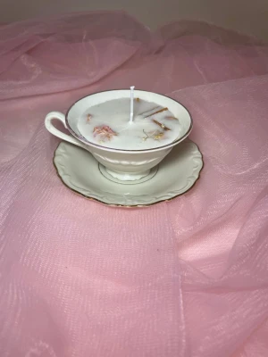 Doftljus i en tekopp i en vit färg med torkade blommor och kanel  - Det här ljuset är gjort av återanvändbara material. Tekoppen är köpt secondhand och stearinet är från kyrkor som tänkte slänga det. Det här ljuset har en svag doft utav vanilj, så känsliga även kan använda det. Det här ljuset har även torkade blommor och kanel på som dekoration. 