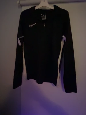 Svart Nike Dri-Fit träningströja barn M - Nike Dri-Fit träningströja i svart med vita detaljer längs sidorna och ärmarna. Tröjan har halv dragkedja framtill och är tillverkad i ett lätt, syntetiskt material som andas. Perfekt för träning eller sportaktiviteter. Tröjan är i väldigt bra skick och storleken är Barn Medium.