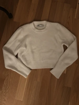 Vit croppad stickad tröja Zara - Säljer en vit croppad stickad tröja från Zara i storlek M. Tröjan har långa ärmar, ribbade muddar och bred ribbad kant nedtill. Perfekt till höst och vinter för en clean och enkel look.