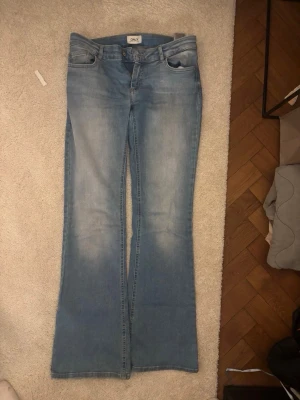 Bootcut jeans från ONLY ljusblå - Säljer ett par ljusblå bootcut jeans från ONLY med klassisk femficksdesign och dragkedja. Jeansen har en snygg tvättad look och är tillverkade i mjukt denimtyg. Perfekta för dig som gillar retrovibbar och vill ha ett par jeans med lite vidare ben.