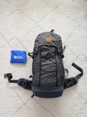 Svart Fjällräven Singi 48 ryggsäck - Stor och robust svart Fjällräven Singi 48 ryggsäck med flera justerbara remmar, vadderade axelband och praktiska fack. Snyggt lädermärke framtill och medföljande blå packpåse. Perfekt för längre äventyr och utflykter.