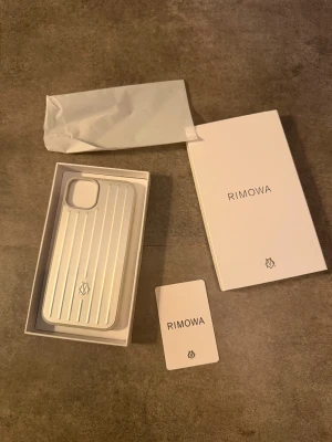 Rimowa iPhone Case - Exklusivt Rimowa iPhone-skal i silver, levereras med originalförpackning och certifikat. Skalets ikoniska räfflade design ger både stil och skydd. Skicket är som nytt, inga synliga repor eller slitage. Perfekt för dig som vill kombinera lyx och funktion till din iPhone.. 14 plus 15 plus 