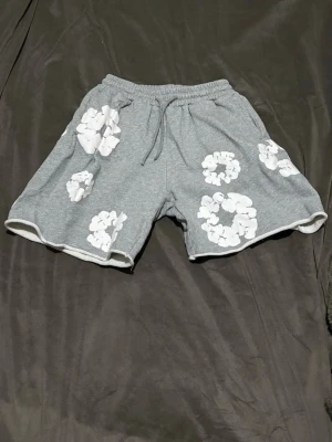 Grå blommiga shorts från Denim Tears - Säljer ett par grå shorts från Denim Tears med vita blommiga tryck över hela plagget. De har elastisk midja med snörning och är tillverkade i mjuk bomull, perfekta för chill dagar. Passformen är loose och de har en avslappnad vibe.