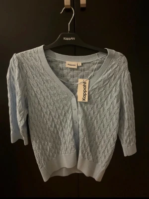 Ljusblå kofta från Kappahl - Supersöt ljusblå kofta från Kappahl med v-ringning och fina stickade detaljer. Koftan har trekvartsärm och knappar framtill. Perfekt till våren och sommaren för en fräsch look. Materialet är mjukt och stickat, vilket gör den både bekväm och snygg.