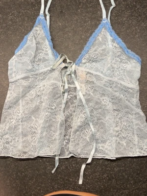 Ljusblå genomskinlig spetslinnetopp med snörning - Säljer en ljusblå, transparent linnetopp i spets med tunna axelband och blå spetskant upptill. Toppen har dekorativ snörning framtill och ett blommigt spetsmönster över hela plagget. Knappt använd