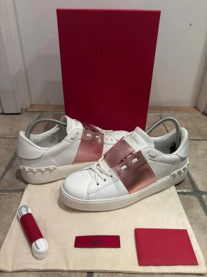 Valentino Garavani sneakers med rosa metallic - Snygga vita Valentino Garavani sneakers med bred metallicrosa rem över vristen och ikoniska nitar på hälen. Skorna är i skinn och har platt sula, rund tå och vita snören. Perfekta för dig som vill sticka ut med en lyxig och trendig look. Storlek 39 i toppen skick, knappt använd sula. Box och dustbag ingår