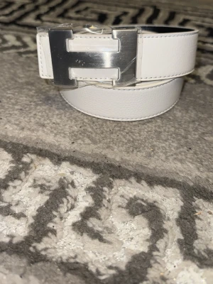 Vit skärp med stor H- silverspänne - Säljer ett stilrent vitt skärp i skinn med ett stort, blankt silverspänne format som ett H. Skärpet har en clean look och passar perfekt till jeans eller byxor för att lyfta outfiten.