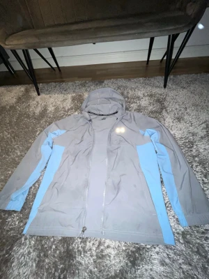 Grå-blå vindjacka från Under Armour - Säljer en grå vindjacka från Under Armour med ljusblå detaljer längs sidorna och ärmarna. Jackan har huva, hel dragkedja framtill och reflekterande logga på bröstet. Perfekt för utomhusaktiviteter när det blåser. Materialet är lätt och syntetiskt.