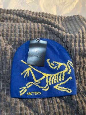 Blå mössa från Arc'teryx med gul logga - Snygg blå mössa från Arc'teryx med deras ikoniska gula fossil-logga framtill. Tillverkad i 100% akryl för en mjuk och bekväm känsla. Perfekt accessoar för att lyfta din look under kyliga dagar.