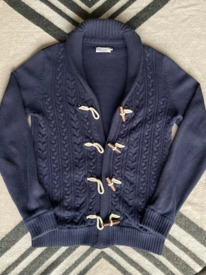 Mörkblå kofta från Pier One med snörning - Säljer en mörkblå kofta från Pier One med grovstickat flätmönster framtill. Koftan har snygga snörningar med träknappar och ribbade muddar. Perfekt för kyliga dagar och ger en riktigt mysig vibe.