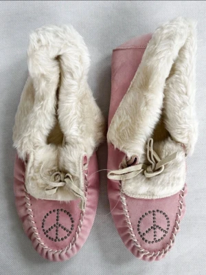 Rosa mockasiner från PINK med pälsfoder - Mysiga rosa mockasiner från PINK med mjukt vitt fuskpälsfoder och snörning framtill. Dekorerade med ett peace-märke i nitar på tån och har en platt sula. Perfekta för kalla dagar när du vill ha det extra cozy. Storleken är L men passar mig som brukar ha 38/39
