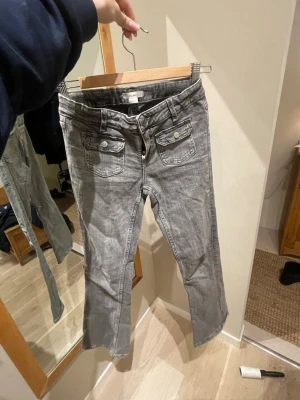 Grå jeans från Gina Tricot, storlek 158 - Säljer ett par grå jeans från Gina Tricot i storlek 158. Byxorna är utsvängda, två framfickor med lock och knapp, samt dragkedja och knapp i midjan. Klassisk jeanslook med bälteshällor och snygg tvättad finish.