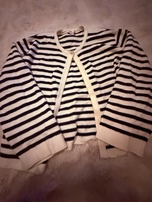 Randig kofta från Gina Tricot - Säljer en snygg randig kofta från Gina Tricot i vitt och marinblått. Koftan har lång ärm, rund halsringning och stängs med en knapp framtill. Materialet är mjukt och skönt, perfekt för lager på lager under höst och vår.