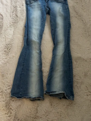 Blå bootcut jeans Gina Tricot M - Säljer ett par blå bootcut jeans från Gina Tricot i storlek M. Jeansen har en ljus tvätt med slitningar längs benen och klassiska bakfickor. Tillverkade i jeansmaterial med snyggt utsvängda ben för en retro vibe.