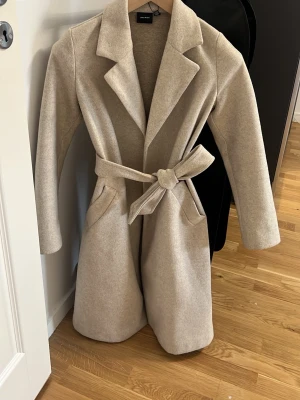 Beige kappa - Beige kappa från Vero Moda med klassisk krage och knytskärp i midjan. Jackan har två fickor och är 100% polyester. Köpte för ca 800, bra kvalite och varm 💞