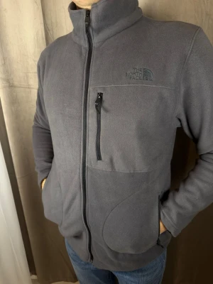 Grå fleecejacka från The North Face - Mjuk och varm grå fleecejacka från The North Face med hel dragkedja, hög krage och tre fickor med dragkedja. Perfekt för kyliga dagar och enkel att slänga på över en t-shirt. Diskret logga på bröstet och stilren design.