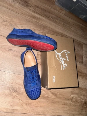 Blå mocka sneakers från Louboutin - Säljer ett par riktigt snygga blå sneakers från Christian Louboutin i mocka med matchande blå snörning och coola nitar på tån. Skorna har den ikoniska röda sulan och en clean, rund tå. Perfekta för dig som vill sticka ut med exklusiv stil, en spik har ramlat av men har den kvar ifall ni vill försöka limma på den, sulorna ser bättre ut i verkligheten än på kamera, priset är ej hugget i sten, ifall ni har några frågor tveka inte på att fråga 