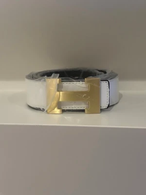 Vit bälte med guldspänne - Snyggt vitt bälte med ett stort, fyrkantigt guldspänne som verkligen sticker ut. Bältet har en clean look och är gjort i ett slätt material med flera hål för justering. Perfekt accessoar för att lyfta vilken outfit som helst.