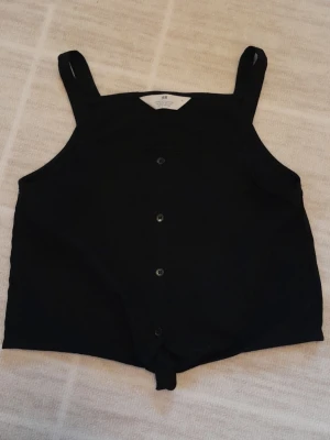 Svart linnetopp med knut - Snygg svart linnetopp från H&M i storlek 152. Toppen har breda axelband, knappar framtill och en knytning längst ner för en trendig look. Perfekt till sommaren och enkel att matcha med olika outfits. !!endast swish +frakt!!