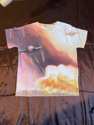 Star Wars T-shirt med Tie Fighter - Cool Star Wars t-shirt med Tie Fighter-tryck på framsidan och laserskott i grönt. Framsidan har en färgstark design med moln och rymdtema, medan baksidan är helvit. Tillverkad i 100% polyester och har klassisk passform med korta ärmar.