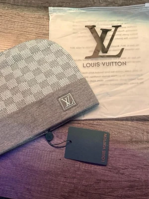 Grå rutig mössa - Snygg grå mössa med klassiskt rutigt mönster och logga framtill. Mössan har en bred uppvikt kant och är tillverkad i mjukt material som känns lyxigt och stilrent. Perfekt accessoar för att lyfta din outfit.