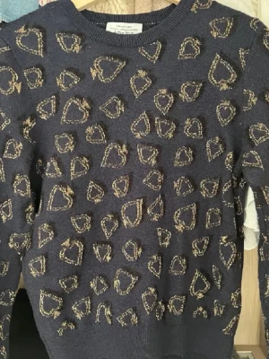 Mörkblå stickad tröja med hjärtan - Mysig mörkblå stickad tröja från & Other Stories Paris Atelier, dekorerad med glittriga guldhjärtan över hela plagget. Rund halsringning och långa ärmar. Perfekt för dig som vill ha något extra i garderoben.