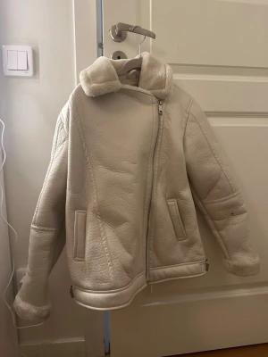 Vit teddyjacka med dragkedja - Säljer en supermjuk och fluffig vit teddyjacka med bred krage och sned dragkedja. Jackan har två fickor framtill och mysigt foder som syns vid ärmsluten. Perfekt för kalla dagar och riktigt snygg till jeans eller kjol.