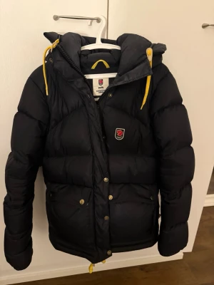 Svart dunjacka Fjällräven Expedition S - Säljer en svart dunjacka från Fjällräven Expedition Series, dammodell i storlek S. Jackan har huva, gula snören och tryckknappar framtill. Perfekt för kalla dagar!