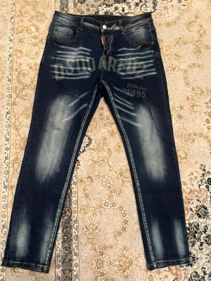  Dsquared2  - Snygga mörkblå jeans från Dsquared2 med blekta detaljer och tydliga Dsquared2-tryck både fram och bak. Klassisk femficksmodell med raka ben och orange kontrastsömmar. Jeansen är i denim och har knappgylf samt läderpatch bak i midjan.