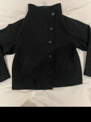 Svart kappa med hög krage - Säljer en svart kappa med rak passform och hög krage. Jackan har fem knappar framtill, diskreta fickor på sidorna och långa ärmar. Enkel och stilren design som passar perfekt till kyligare dagar.